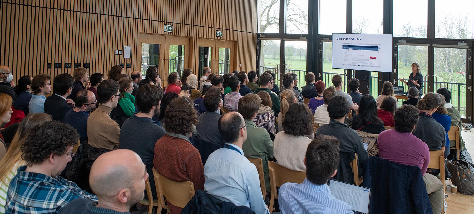 A room of people looking at a presentation screen. The room has a wall-to ceiling window behind the screen.