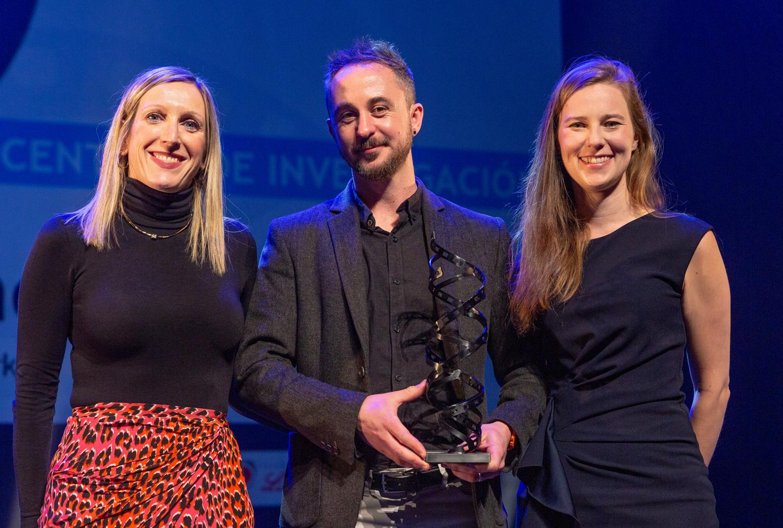 A woman, a man and another woman stand facing the camera, the man in the middle holds a metal award- a sculpture of two film strips in a double helix.